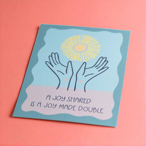Uplifting A Joy Shared Is A Joy Made Double print in blue, pink and yellow, styled against a bright coral pink background.