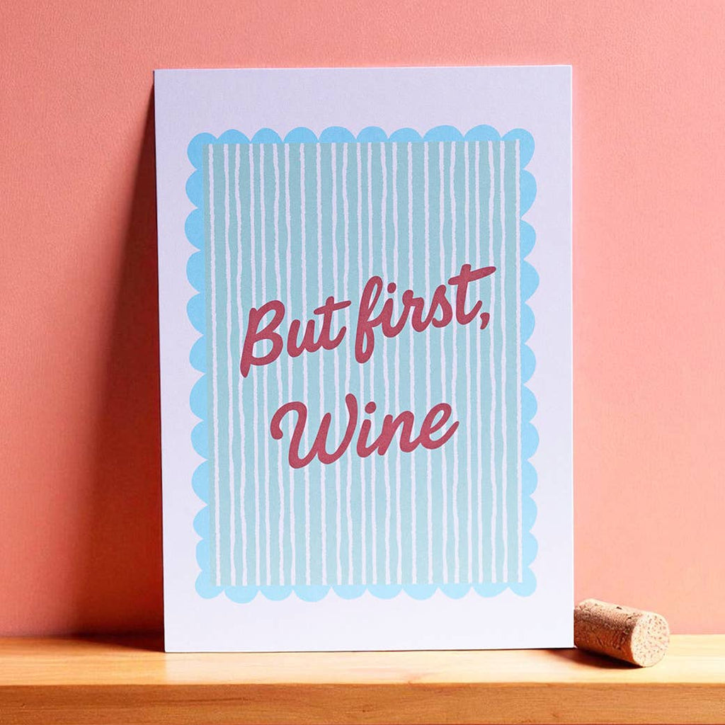 Cheerful But First Wine A5 print with red script lettering and aqua stripes, displayed against a warm coral pink wall.