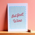 Cheerful But First Wine A5 print with red script lettering and aqua stripes, displayed against a warm coral pink wall.