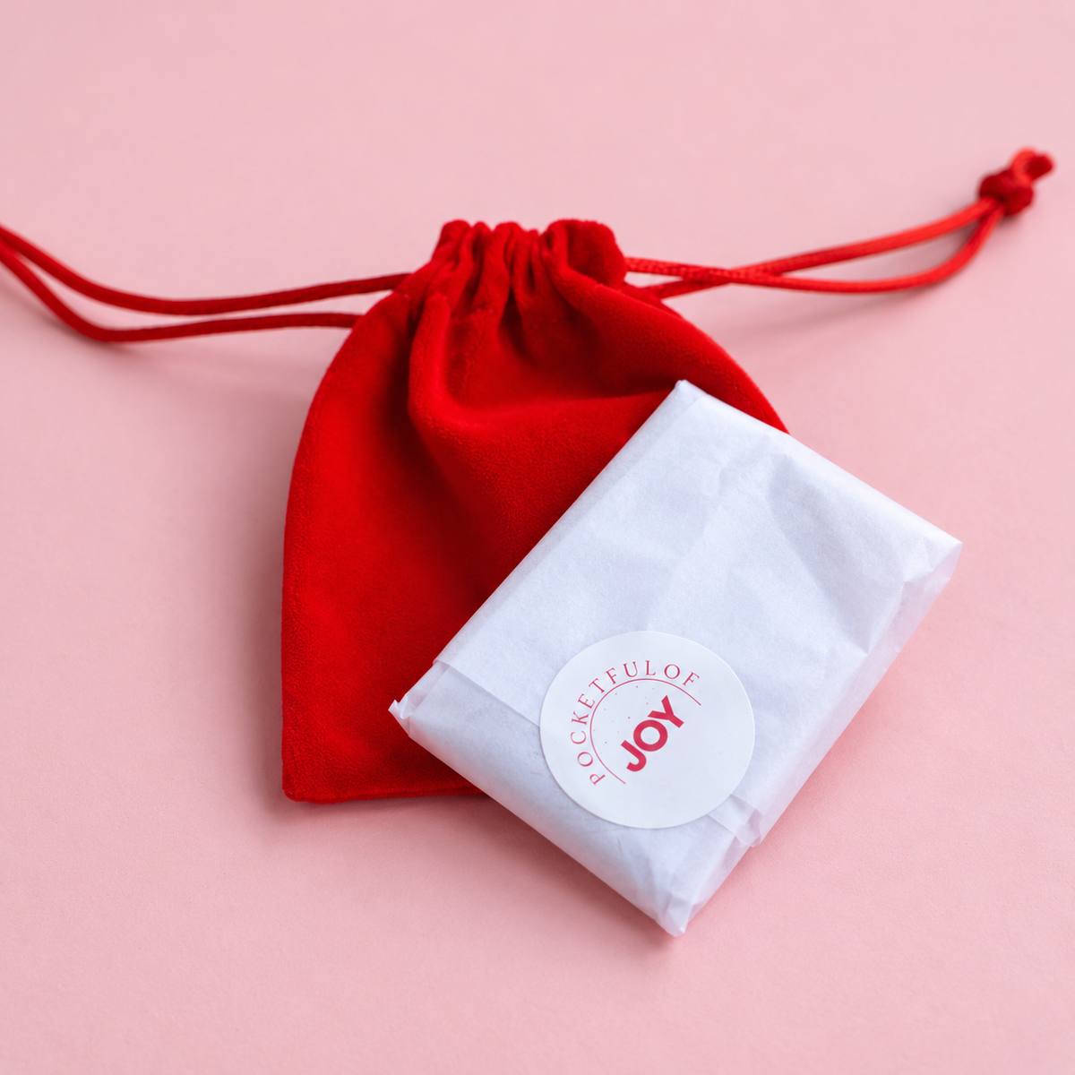 A small white tissue-wrapped package with a round white sticker reading 'Grateful for JOY' placed in front of a red drawstring pouch on a soft pink background.