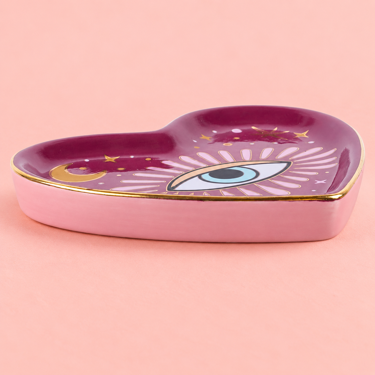 A heart-shaped pink ceramic dish with a glossy deep magenta top, decorated with a celestial eye motif, gold stars, and a gold crescent moon, set against a solid pink background.