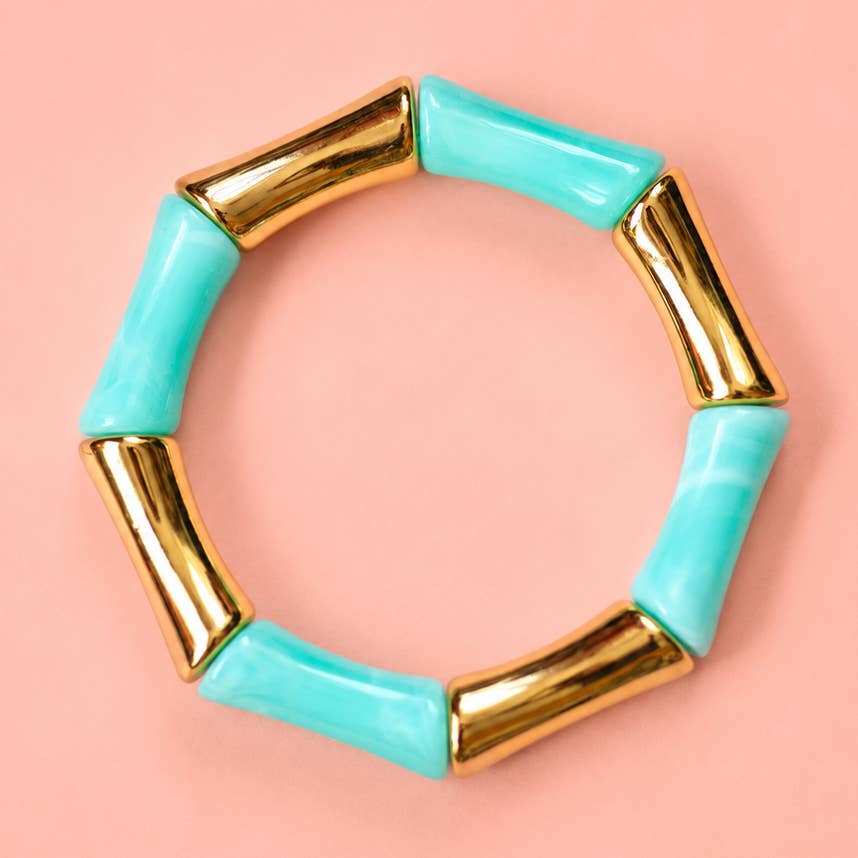 Aqua acrylic and polished gold tone bamboo inspired stretch bracelet on a pink background.