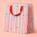 Pale pink gift bag with colourful vertical stripes, tiny bow details and a bright red ribbon handle.
