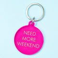 A bright pink circular keychain tag with the phrase 'NEED MORE WEEKEND' in gold lettering, attached to a silver metal key ring, set against a light blue background.