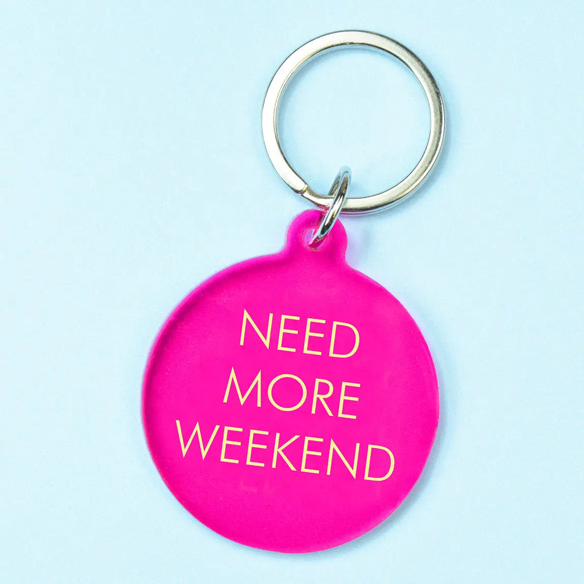 A bright pink circular keychain tag with the phrase 'NEED MORE WEEKEND' in gold lettering, attached to a silver metal key ring, set against a light blue background.
