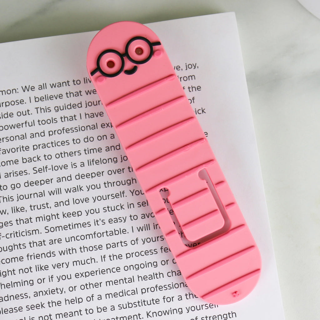 Pink bookworm reading light and bookmark with black glasses resting on an open book.
