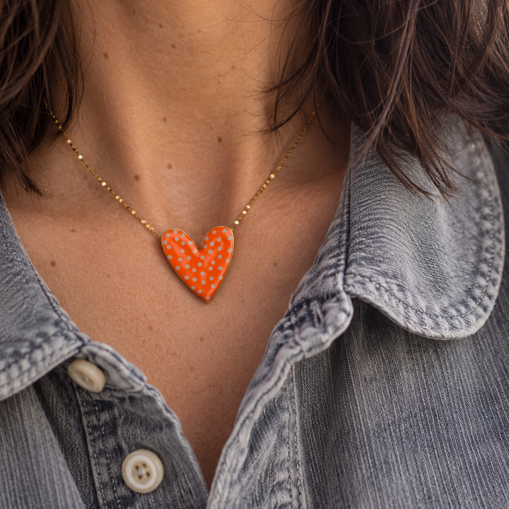 Woman wearing an orange and white polka dot enamel heart necklace with a grey denim shirt.