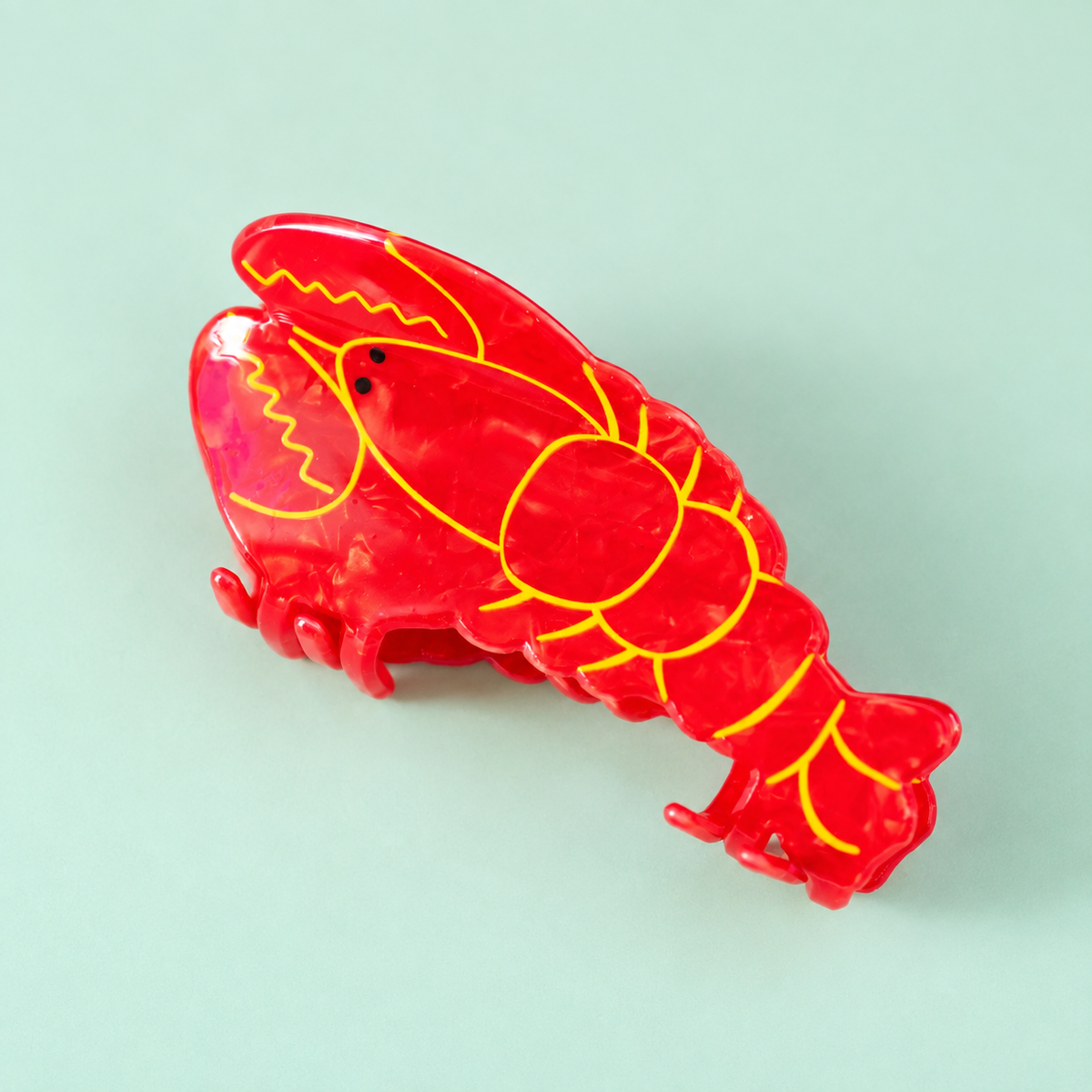 Bright red lobster shaped hair claw clip with yellow detailing and black eyes on a soft pale mint background.