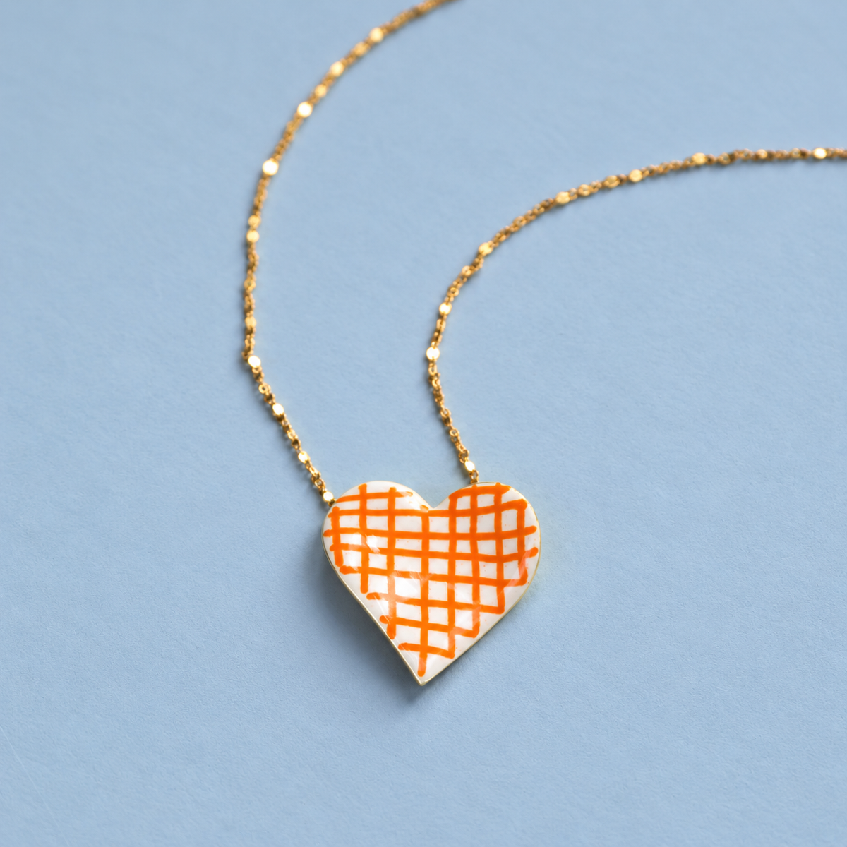 Limited edition gold plated heart necklace with cream enamel and a bold orange check pattern on a soft pale blue background.