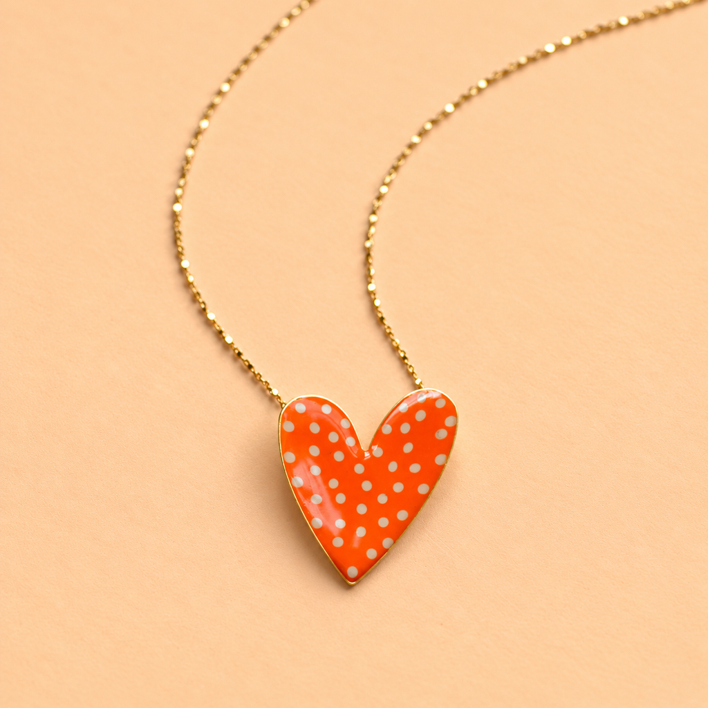 Gold plated heart necklace with bright orange enamel and white polka dots on a soft peach background.