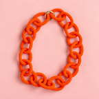 Chunky orange beaded statement chain necklace with oversized interlocking links and gold tone ring fastening on a soft pink background.
