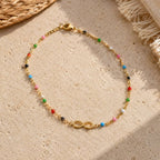 Delicate gold infinity anklet with colourful beads and adjustable clasp laid on a neutral stone surface.
