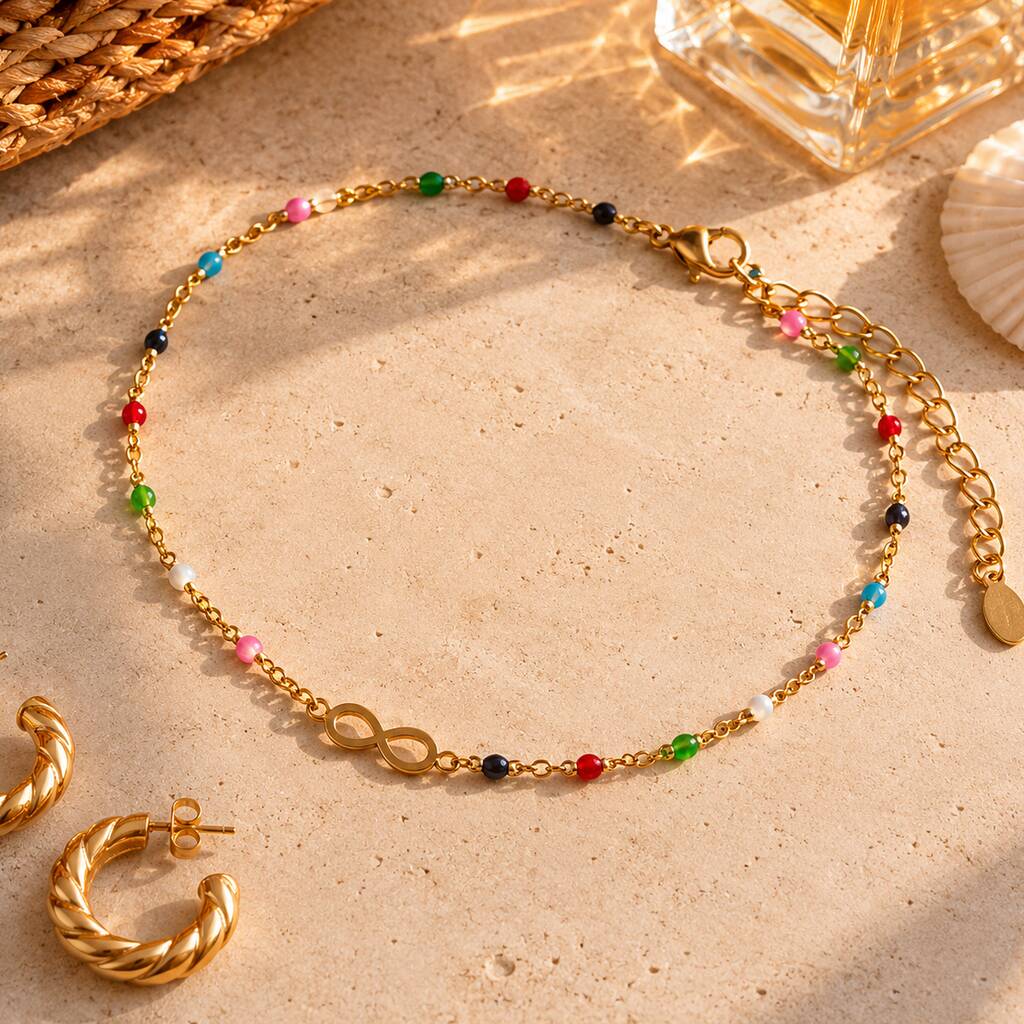 Gold tone infinity anklet with tiny multicoloured beads displayed on a neutral stone background.
