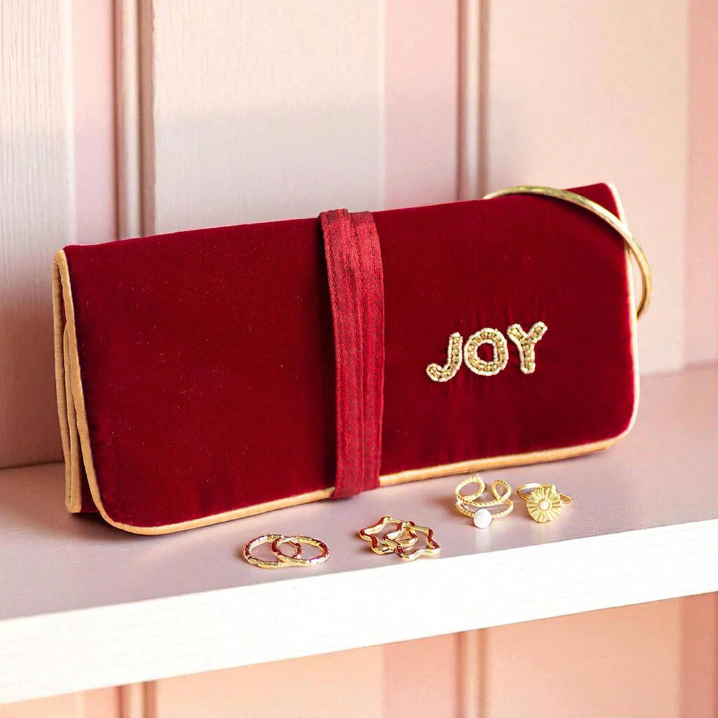Red velvet JOY travel jewellery pouch with gold jewellery displayed on top, styled with holiday accessories.