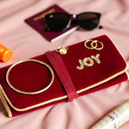 Red velvet travel jewellery pouch with gold piping, sparkling JOY lettering and gold jewellery displayed beside it.