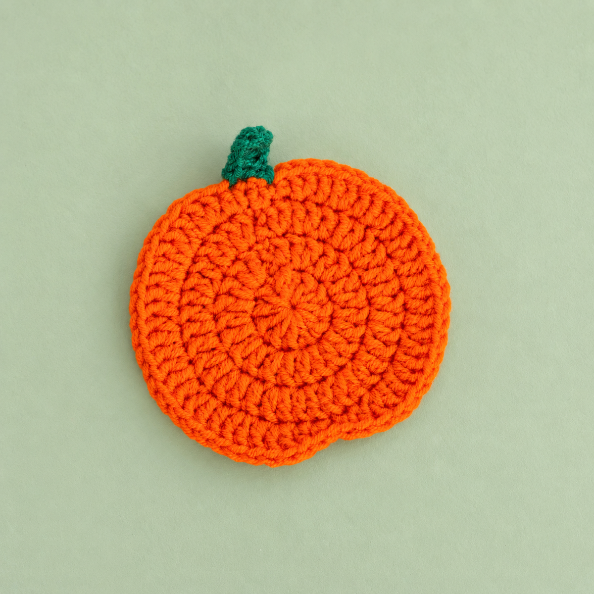 Limited edition handmade orange crochet pumpkin with green stalk on a soft sage background.