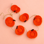 Handmade crochet pumpkin garland with six bright orange pumpkins on a soft peach background.