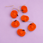 Six orange crochet pumpkins strung on natural twine, photographed on a soft lilac background.