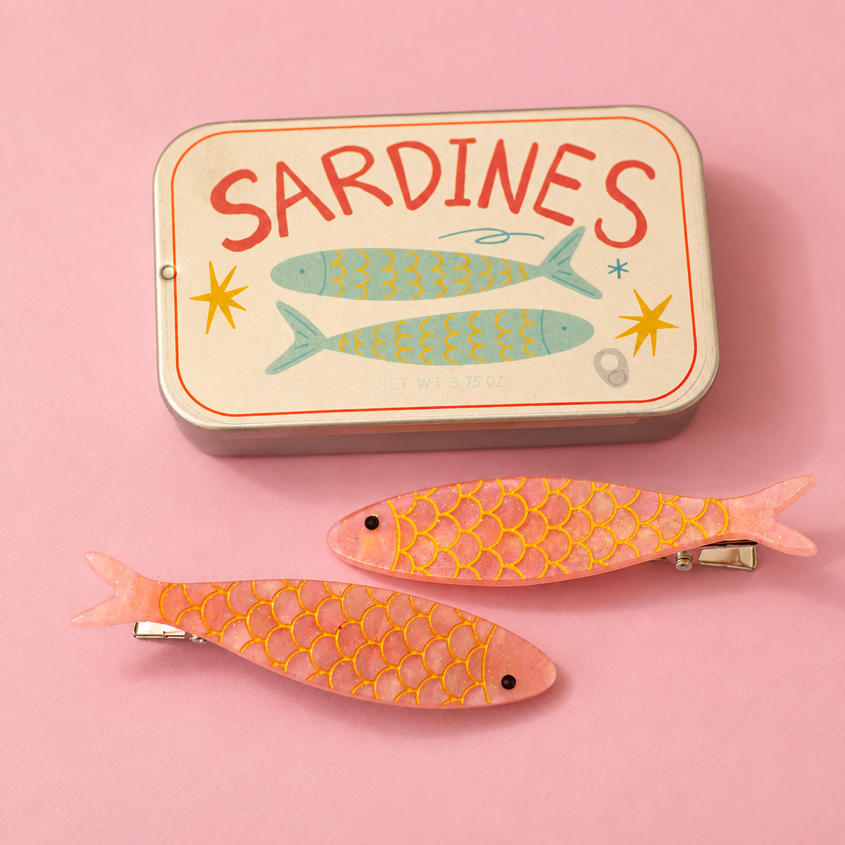 Two pink glitter sardine hair clips with gold scale detailing beside a vintage style sardine tin.