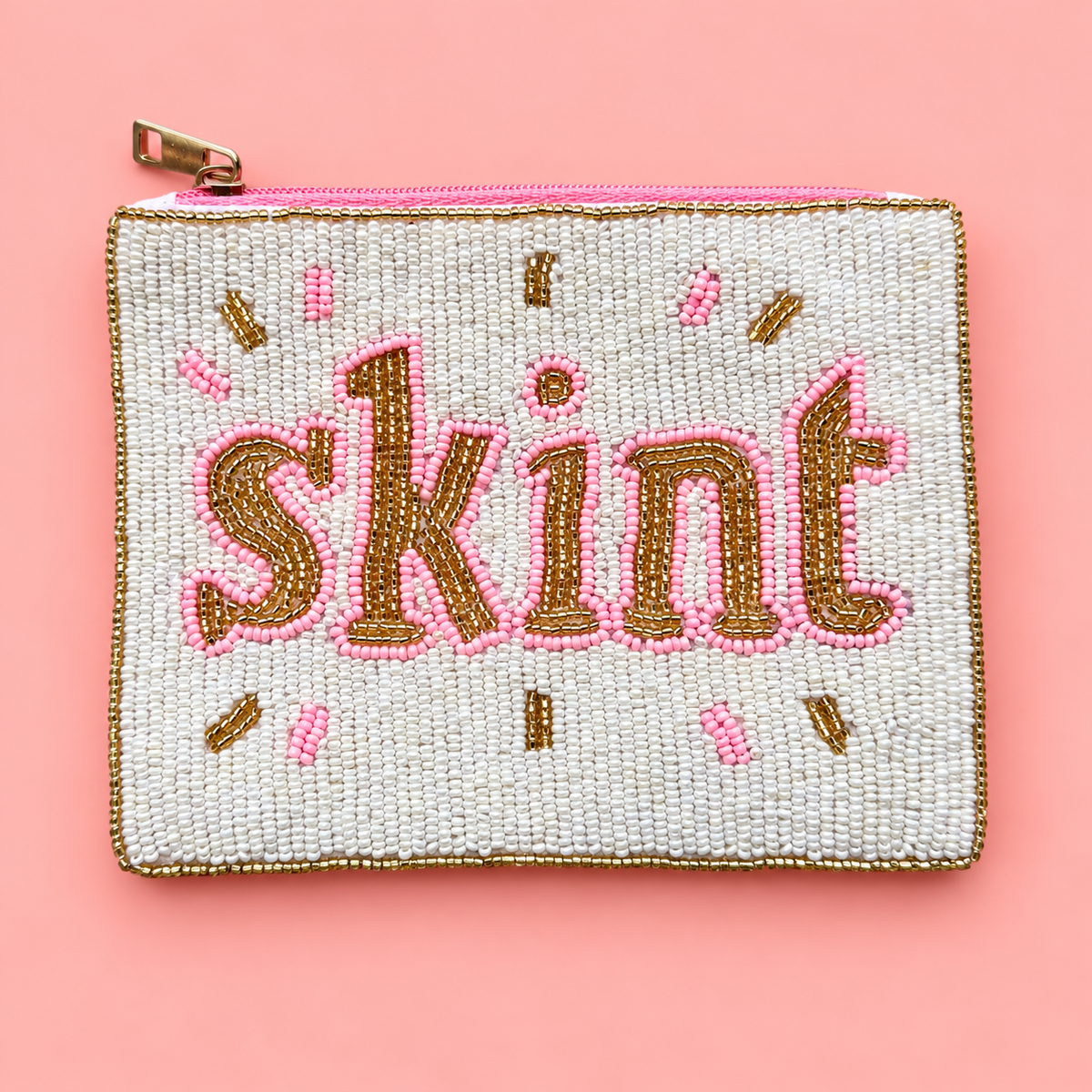 A rectangular beaded coin purse with the word 'skint' in gold and pink lettering, set against a light pink background.
