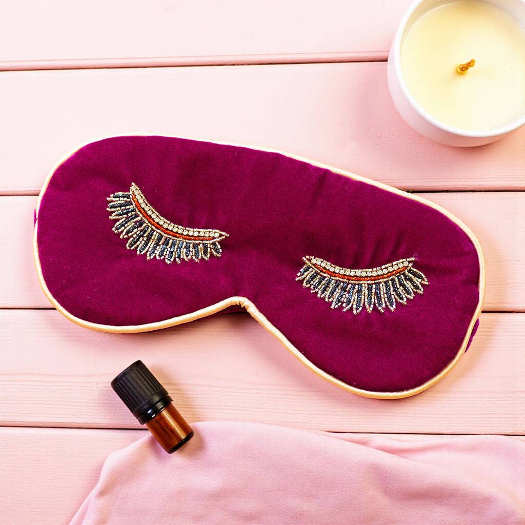 Magenta velvet eye mask with beaded eyelashes and pale gold edging displayed with relaxation accessories on a pink background.