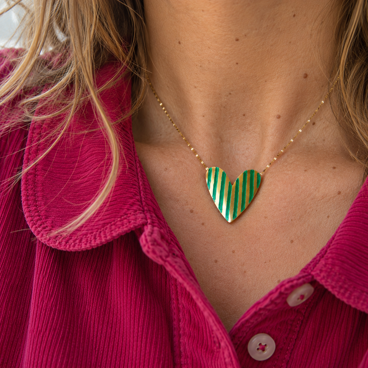A close-up of a person wearing a gold chain necklace with a heart-shaped pendant featuring diagonal green and white stripes, styled with a bright pink corduroy shirt.